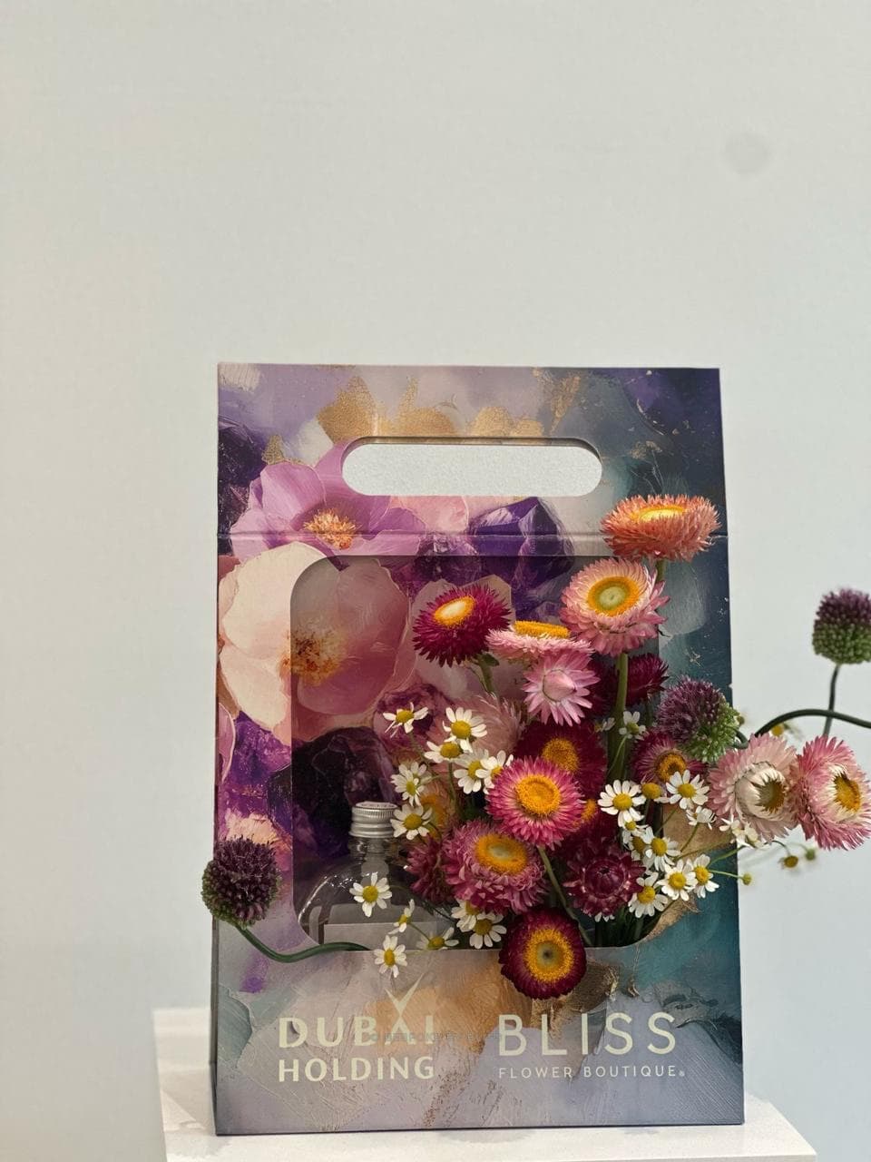 A branded gift carrier in violet and blue tones holding flowers beside a small glass bottle