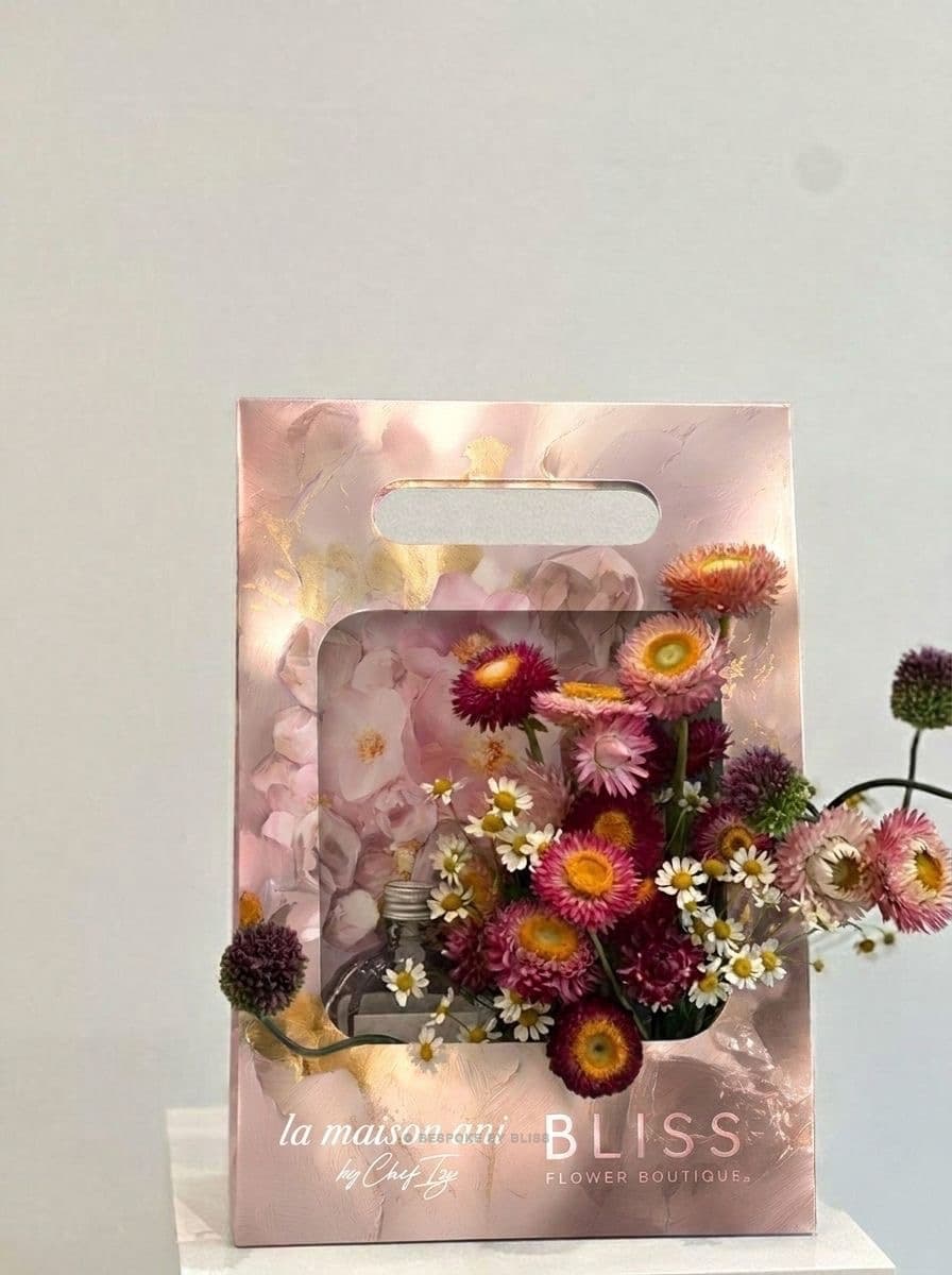 A pink branded gift carrier holding dried and fresh flowers beside a small glass bottle