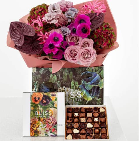 A flower bouquet in pink paper above a branded box and a tray of chocolates